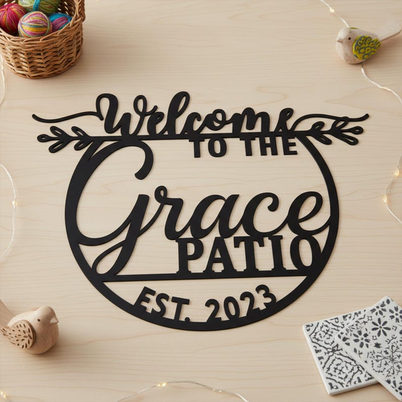 K&ouml;nnte beinhalten: Schwarzes Metall-Terrassenschild mit den Worten "Welcome to the Grace Patio EST. 2023" in einer dekorativen Schriftart. Das Schild ist kreisf&ouml;rmig und hat ein Blattdesign. Das Schild befindet sich auf einer hellen Holzoberfl&auml;che.