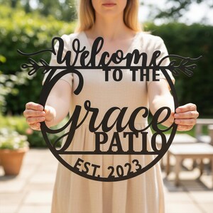 K&ouml;nnte beinhalten: Schwarzes Metallschild mit der Aufschrift "Welcome to the Grace Patio" und "EST. 2023". Das Schild hat eine runde Form mit dekorativen Verzierungen. Eine Person in einem beigen Kleid h&auml;lt das Schild.