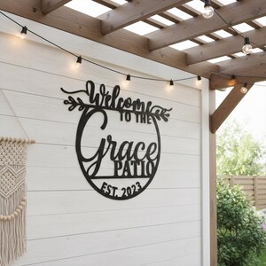 K&ouml;nnte beinhalten: Schwarzes Metallschild mit der Aufschrift "Welcome to the Grace Patio EST. 2023" in dekorativer Schrift. Das Schild ist an einer wei&szlig;en Holzwand befestigt, dar&uuml;ber Lichterketten. Links ist ein Makramee-Wandbehang zu sehen.