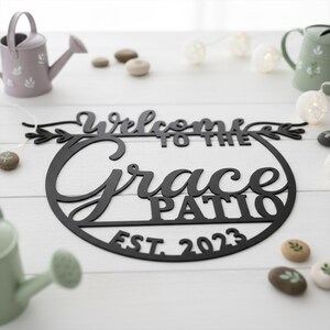 K&ouml;nnte beinhalten: Schwarzes Metallschild mit der Aufschrift "Welcome to the Grace Patio EST. 2023". Das runde Schild hat eine dekorative Schriftart und florale Elemente. Es liegt auf einer wei&szlig;en Holzoberfl&auml;che.