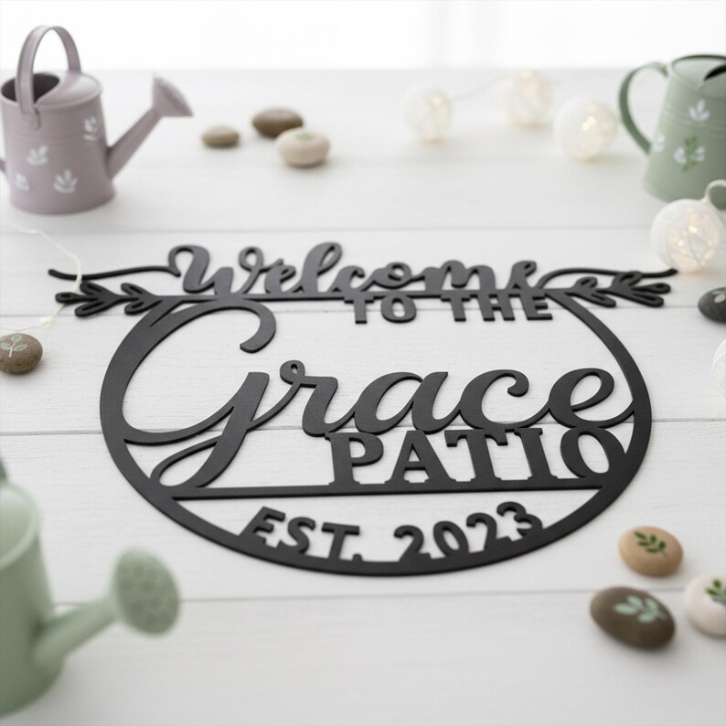 K&ouml;nnte beinhalten: Schwarzes Metallschild mit der Aufschrift "Welcome to the Grace Patio EST. 2023". Das runde Schild hat eine dekorative Schriftart und florale Elemente. Es liegt auf einer wei&szlig;en Holzoberfl&auml;che.