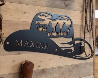 Könnte beinhalten: Schwarze Metall-Wandkunst in Form eines Cowboyhuts, mit einer Silhouette von Reitern auf Pferden und dem Namen "MAXINE". Der Hut ist mit Sternen und einer Berglandschaft verziert. Ein brauner Leder-Cowboystiefel ist darunter zu sehen.