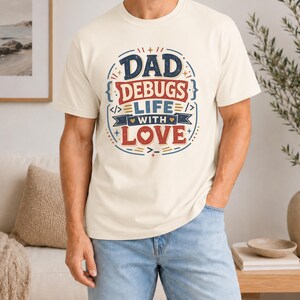 Puede incluir: Camiseta color crema con el texto "DAD DEBUGS LIFE WITH LOVE" en un diseño circular. El diseño presenta detalles en rojo, azul y dorado, y un símbolo de corazón. La camiseta se lleva con vaqueros azules.