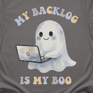 May include: A charcoal grey t-shirt featuring a graphic of a white ghost using a laptop. Above the ghost, the text reads MY BACKLOG in a curved, multi-coloured font. Below the ghost, the text reads IS MY BOO in the same multi-coloured font.
