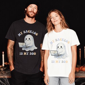 May include: A dark charcoal grey t-shirt and a white t-shirt, each featuring a graphic of a cute ghost using a laptop. The text "MY BACKLOG IS MY BOO" is printed in a colourful, retro-inspired font above and below the ghost illustration.