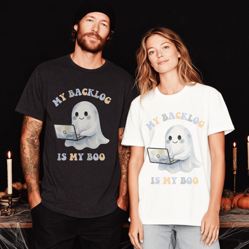 May include: A dark charcoal grey t-shirt and a white t-shirt, each featuring a graphic of a cute ghost using a laptop. The text "MY BACKLOG IS MY BOO" is printed in a colourful, retro-inspired font above and below the ghost illustration.
