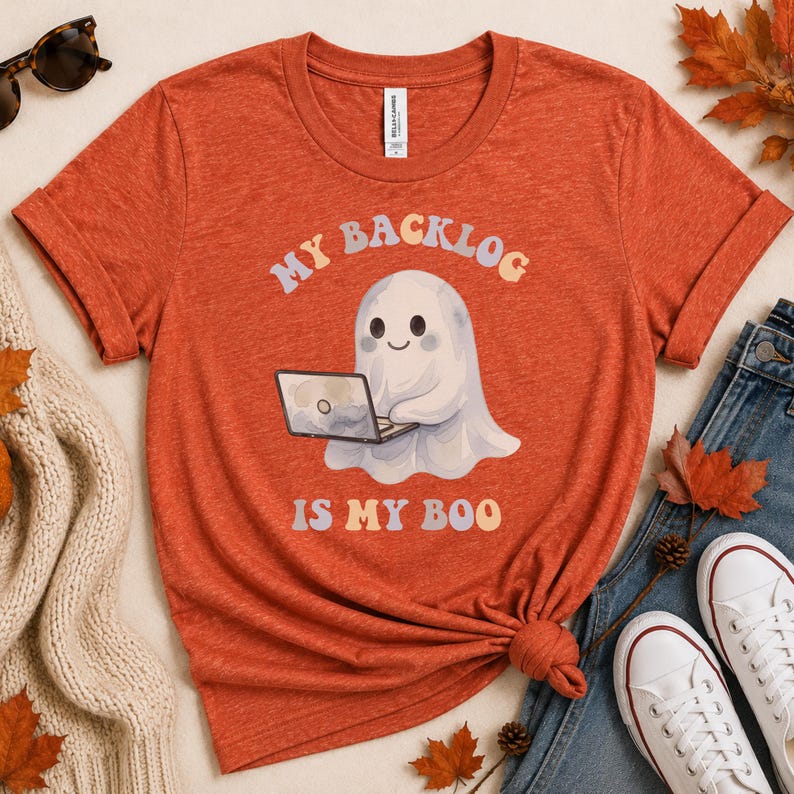 May include: A burnt orange short-sleeve t-shirt featuring a graphic of a white ghost sitting at a laptop. The text above the ghost reads "MY BACKLOG" and the text below reads "IS MY BOO" in a colourful, rounded font. The shirt has a crew neckline.