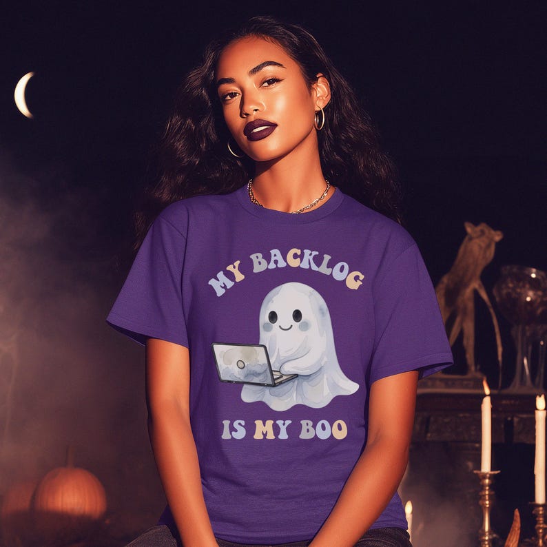 May include: A purple short-sleeved t-shirt featuring a graphic of a white ghost sitting with a laptop. The text above the ghost reads MY BACKLOG and the text below reads IS MY BOO in a light-coloured, wavy font. The shirt has a crew neckline.