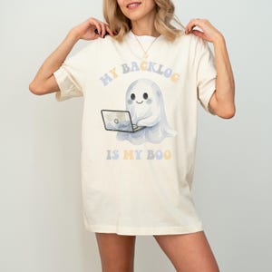May include: An off-white short-sleeve t-shirt featuring a graphic of a white ghost using a laptop. The text above the ghost reads MY BACKLOG and the text below reads IS MY BOO in colourful, rounded, pastel-toned lettering on the front centre.