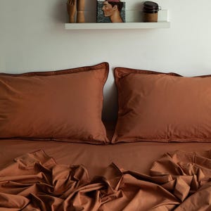 May include: Rust-colored bed linens, including two pillowcases and a fitted sheet, are neatly arranged on a bed. The pillowcases have a darker trim. A shelf above the bed displays decorative items, including a book and a small wooden hand.