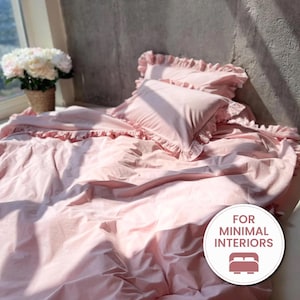 May include: A light pink bedding set featuring a duvet cover and two pillowcases with ruffled edges. The fabric has a smooth texture and is draped over a bed. A circular white badge in the corner displays the text FOR MINIMAL INTERIORS with a bed icon.