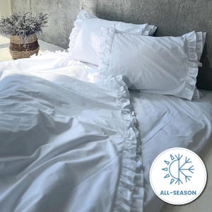 May include: White bedding set with ruffled edges, including two pillowcases and a duvet cover. The all-season bedding is made of soft fabric and is neatly arranged on a bed. A basket of dried flowers is in the background.