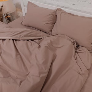 Può includere: Un letto con un copripiumino color rosa antico e due federe abbinate. Le federe e il copripiumino hanno un bordo bianco. La biancheria da letto è disposta su una struttura letto bianca.