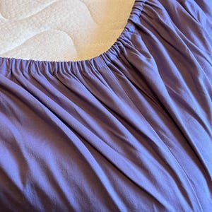 May include: A solid light purple fitted sheet with an elasticated edge, shown partially covering a white mattress. The fabric features a smooth texture with gathered folds along the perimeter where the elastic band secures the sheet in place.