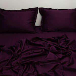 Pode incluir: Um conjunto de lençóis roxos escuros, com duas fronhas e um lençol com elástico. O tecido é drapeado e dobrado, criando sombras suaves. A roupa de cama é de cor sólida, oferecendo uma estética simples e elegante.