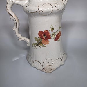 May include: Cream-colored ceramic pitcher with a decorative handle and spout. The pitcher features a floral design of red poppies and green leaves. Gold-colored accents adorn the rim and base, adding a touch of elegance to the vintage-style piece.