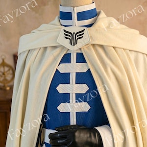 May include: A cream-colored cloak and blue tunic with white accents. The tunic features a series of white, horizontal, rectangular shapes. A black emblem is on the cloak, and black gloves are visible.
