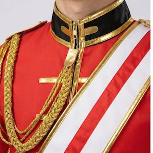 Peut inclure: Veste rouge de style militaire avec des bordures dorées, un col noir et une écharpe blanche et rouge. La veste présente des épaulettes tressées dorées et une fermeture éclair dorée. Le design est formel et orné.