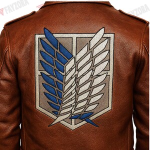 K&ouml;nnte beinhalten: Braune Lederjacke mit einem gro&szlig;en, gestickten Emblem auf dem R&uuml;cken. Das Emblem zeigt blaue und wei&szlig;e Fl&uuml;gel in einem rechteckigen Rahmen. Die Jacke hat ein klassisches Design mit Schulterklappen.