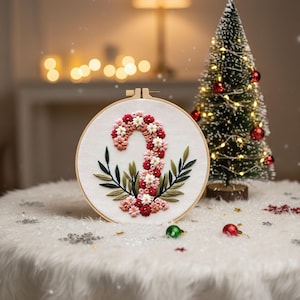May include: Embroidered Christmas candy cane design in a wooden hoop. The candy cane is made of red, pink, and white flowers, with green leaves. A small Christmas tree with lights and ornaments is in the background.