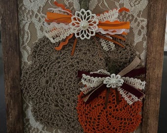 May include: A framed autumn-themed artwork featuring two crocheted pumpkins. The larger pumpkin is brown, and the smaller one is orange. Both are adorned with decorative ribbons and a floral embellishment. The background is a cream-colored lace pattern, all within a wooden frame.