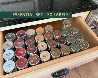 May include: A set of 30 glass spice jars with silver lids, organized in a wooden drawer. Each jar features a white label with black text identifying various spices and seasonings. The text ESSENTIAL SET - 30 LABELS is displayed at the top.