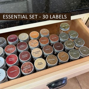 Op de afbeelding: Een houten lade gevuld met kleine glazen kruidenpotjes met zilveren deksels. Elk potje heeft een label met de kruidennaam. De tekst "ESSENTIAL SET - 30 LABELS" staat bovenaan. Kruiden zijn paprika, kaneel en knoflook.