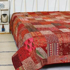 May include: A patchwork quilt in shades of red, orange, and pink covers a bed. The quilt is made of various fabric squares with different patterns and textures. The bed frame is white, and the quilt's edge is a solid red.