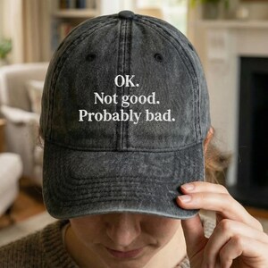 May include: A dark gray baseball cap with the text "OK. Not good. Probably bad." in white. The cap has a curved brim and a distressed, vintage look. The cap is being worn by a person.
