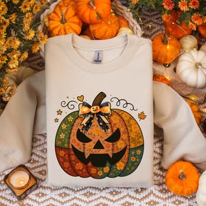 Può includere: Felpa color crema con un design patchwork di una zucca di Halloween. La zucca ha una faccia intagliata, un fiocco e motivi floreali. L'immagine include zucche e foglie autunnali.