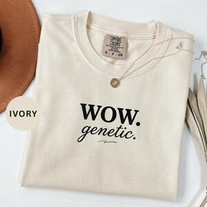 以下が含まれることがあります： 「WOW. genetic.」の黒い文字がプリントされたアイボリー色のTシャツ。クルーネックで、背面上部に小さなタグが付いています。ペンダント付きのシルバーネックレスがシャツの上にかけられています。