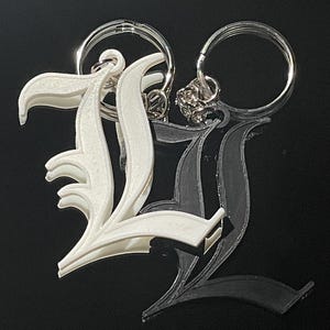 Puede incluir: Dos llaveros, cada uno con la letra mayúscula "L" en fuente gótica. Un llavero es blanco, el otro es negro. Ambos tienen llaveros plateados.