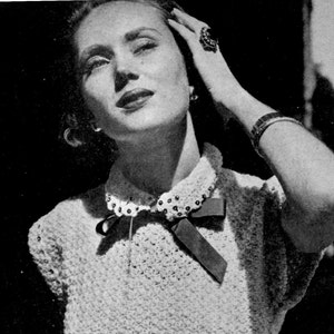 May include: A close-up of a woman wearing a white crocheted sweater with a black bow and pearl embellishments. She is wearing a ring on her finger and a bracelet on her wrist.