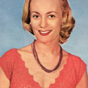May include: A vintage portrait of a woman with blonde hair, wearing a coral-coloured top and a layered pearl necklace. The woman is smiling, with red lipstick, against a light blue background. The top has a scalloped neckline.