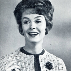 May include: A woman with a bouffant hairstyle smiles while wearing a light-colored crocheted cardigan with a dark floral brooch.