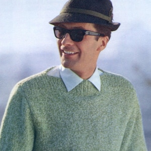 May include: A man wearing a green sweater, a white collared shirt, and a black fedora with a brown band. He is wearing sunglasses.