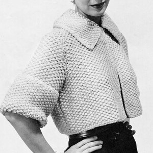 May include: A woman wearing a white knitted cardigan with a large collar and a black skirt. She is wearing a bracelet on her wrist.
