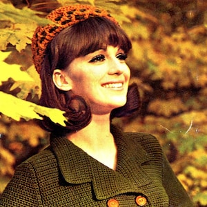 May include: A woman with brown hair wearing a green crocheted jacket and a brown crocheted hat. She is smiling and looking to the right.