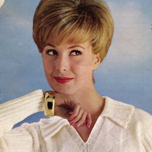 Puede incluir: Imagen vintage de una mujer con cabello rubio y l&aacute;piz labial rojo. Lleva un su&eacute;ter color crema con cuello y una pulsera dorada con un acento negro. El fondo es un cielo azul claro.