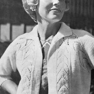 Puede incluir: Una mujer lleva un cárdigan de ganchillo blanco con un patrón de encaje. Lleva una camisa estampada debajo y un cinturón alrededor de la cintura. La foto está en blanco y negro.