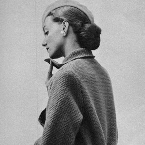 Op de afbeelding: Een vrouw draagt een grijs gebreid jasje met een witte hoed. Ze kijkt naar beneden en heeft haar hand op haar kin.