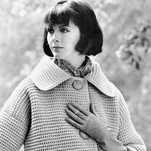 May include: A close-up of a woman wearing a crocheted cardigan with a large collar and a single button. She is wearing a patterned scarf and gloves. The cardigan is a light beige colour.