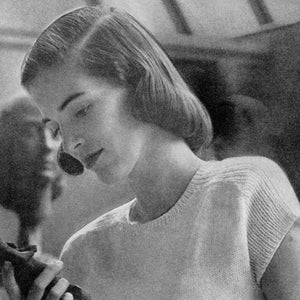 May include: A woman with short, straight hair is looking down at a small, dark object in her hands. She is wearing a white, knitted sweater with short sleeves.