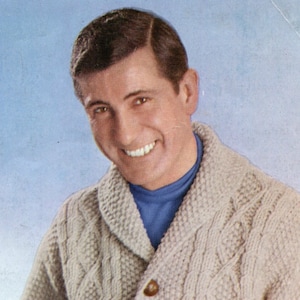 May include: A man with brown hair is smiling and wearing a light brown cable knit cardigan with a blue turtleneck underneath.