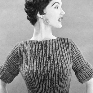 May include: A black and white photo of a woman wearing a knitted sweater with short sleeves. The sweater has a ribbed texture and a round neckline. The woman is wearing earrings and has her hair styled in a short, curly bob.