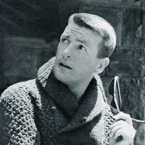 May include: A black and white photo of a man with short, dark hair. He is wearing a knitted sweater with a high collar and is looking up and to the right. He is holding a pair of sunglasses in his right hand.
