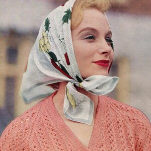 May include: A woman wearing a pink knitted cardigan with a white headscarf with a floral pattern. The headscarf is tied in a knot at the back of her neck.