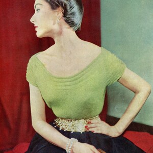 May include: A woman wearing a green knitted sweater with a pearl belt and a black skirt. She is sitting on a red and green surface.
