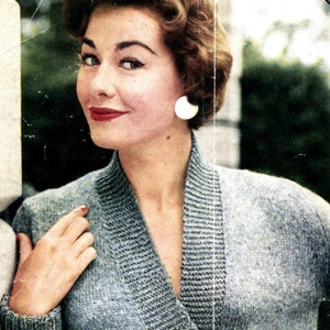 May include: A woman with short, curly brown hair is wearing a grey knitted cardigan with a V-neck. She is wearing a white earring in her left ear.