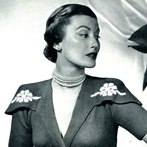 Puede incluir: Una foto en blanco y negro de una mujer con un suéter de punto con cuello en V y adornos florales en los hombros. Lleva un collar de perlas.